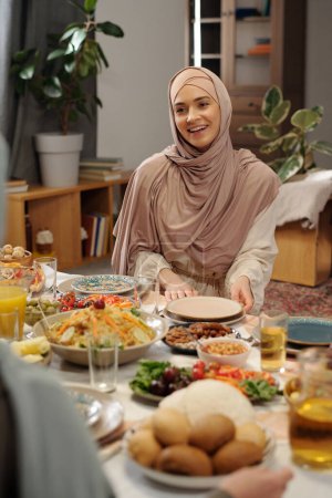 Téléchargez les photos : Plan vertical d'une jeune femme musulmane joyeuse portant le hijab assise à une table festive parlant à sa famille le jour d'Uraza Bayram - en image libre de droit