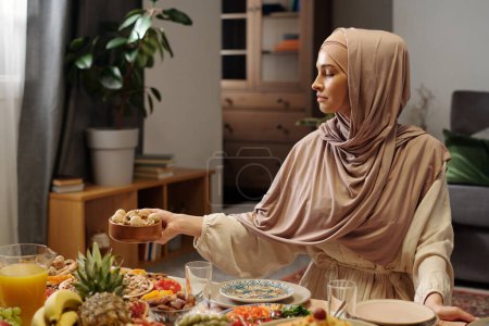 Foto de Foto mediana de una joven musulmana con hijab sentada en una mesa festiva colocando un cuenco de madera lleno de nueces - Imagen libre de derechos