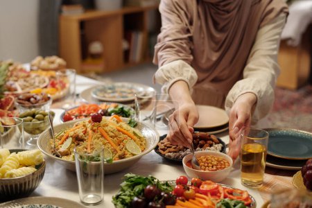 Unrecognizable young Muslim woman wearing hijab setting festive table putting spoon into chickpea dish, copy space