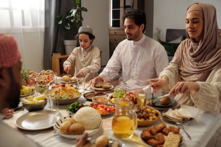 Foto de Familia musulmana moderna reunida en la mesa festiva en la sala de estar comiendo cena casera en Eid Al-Fitr - Imagen libre de derechos