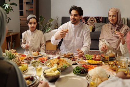 Foto de Moderna familia musulmana con ropa modesta sentada a la mesa en la sala de estar disfrutando de una cena festiva en Eid Al-Fitr - Imagen libre de derechos
