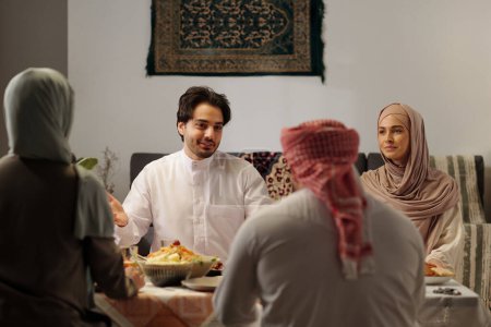 Foto de Captura selectiva de enfoque del joven musulmán sentado en la mesa contando historias durante la cena familiar en Eid Al-Fitr - Imagen libre de derechos