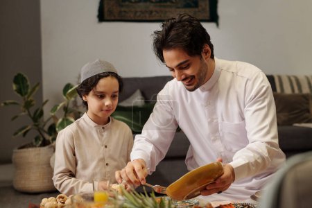 Foto de Mediana toma de alegre joven musulmán usando kandora compartir plato con su hijo durante la cena familiar en sala majilis - Imagen libre de derechos