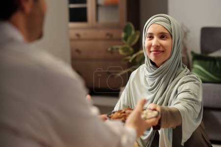 Foto de Sobre el hombro toma de alegre mujer musulmana usando hijab pasando plato con nueces a su pariente masculino durante la cena familiar - Imagen libre de derechos