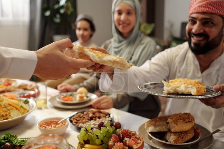 Foto de Captura selectiva de foco de alegre familia musulmana compartiendo pan mientras cenan festivamente en Eid Al-Fitr - Imagen libre de derechos
