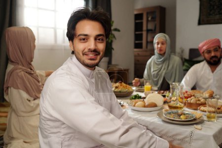 Foto de Retrato de un joven musulmán feliz usando kandora sentado en la mesa festiva con su familia en Eid Al-Fitr sonriendo a la cámara - Imagen libre de derechos