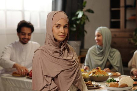 Foto de Retrato de una joven musulmana con hiyab sentado contra una mesa festiva mirando a la cámara en la cena de celebración de Eid Al-Fitr - Imagen libre de derechos
