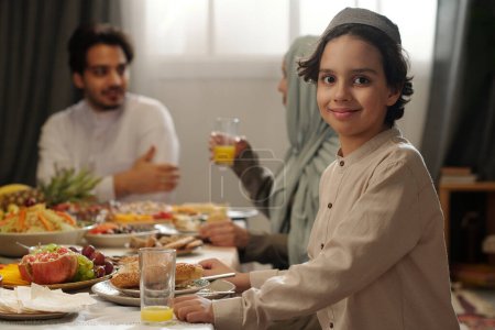 Foto de Retrato de enfoque selectivo de niño musulmán feliz sentado en la mesa festiva mirando a la cámara mientras tiene la cena de celebración de Uraza Bayram con la familia - Imagen libre de derechos