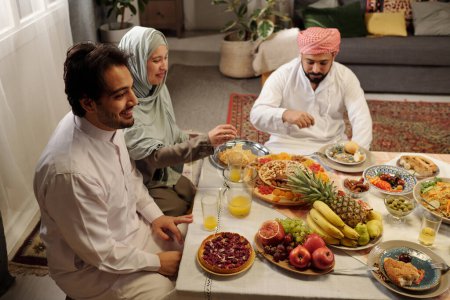 Aufnahme aus der Vogelperspektive von fröhlichen muslimischen Männern und Frauen, die am Tisch im Wohnzimmer sitzen und Uraza Bayram mit ihrer Familie feiern