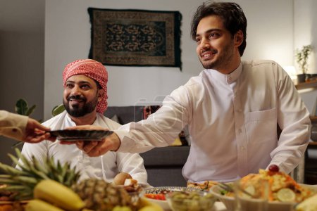 Téléchargez les photos : Plan moyen d'un jeune homme musulman joyeux passant un plat à une femme méconnaissable lors d'un dîner festif en famille à l'Aïd Al-Fitr - en image libre de droit