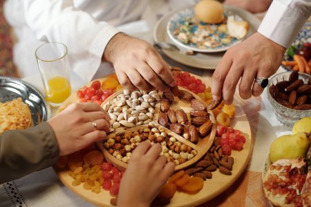 Téléchargez les photos : Mains de personnes musulmanes méconnaissables prenant des noix et des fruits secs du plateau lors du dîner de fête islamique - en image libre de droit