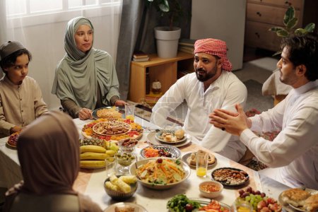 Blick aus der Vogelperspektive auf die moderne muslimische Familie, die Eid Al-Fitr feiert, sitzt am üppigen Festtisch und genießt das Gespräch