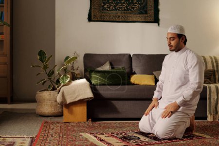 Téléchargez les photos : Jeune musulman du Moyen-Orient assis sur un tapis de prière avec les yeux fermés faire namaz dans le salon à la maison - en image libre de droit