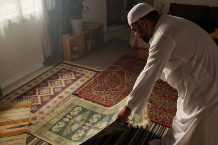 Foto de Vista de ángulo alto del hombre musulmán que pone alfombra de oración verde en el suelo en la sala de estar antes de hacer namaz, espacio de copia - Imagen libre de derechos