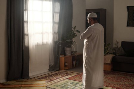 Foto de Hombre musulmán vistiendo thobe de pie en la alfombra de oración en la sala de estar en frente de la ventana haciendo namaz, espacio de copia - Imagen libre de derechos