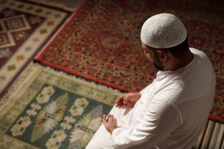 Foto de Vista de alto ángulo de tiro de hombre musulmán maduro con ropa blanca con taqiyah sentado en la alfombra de oración haciendo namaz, espacio de copia - Imagen libre de derechos