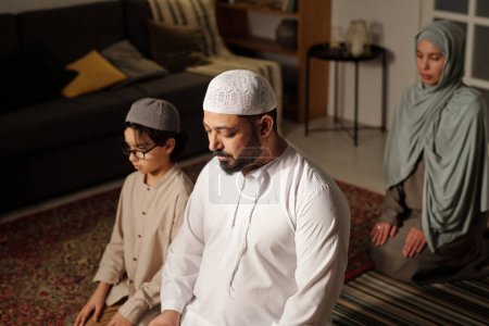 Selektive Fokusaufnahme des modernen muslimischen Mannes, seiner Frau und seines Sohnes, die zusammen im Wohnzimmer sitzen und Salah beten