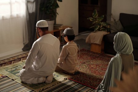Blick aus der Vogelperspektive auf eine nicht erkennbare muslimische Familie mit Kind, das auf dem Boden im Wohnzimmer sitzt und Salah betet