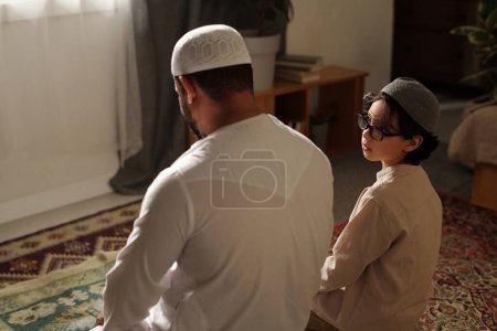 Muslimischer Vater und Sohn tragen Takiya und sitzen auf Teppichen auf dem Fußboden im Wohnzimmer und beten Salah