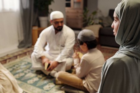 Téléchargez les photos : Mise au point sélective plan grand angle de la jeune femme musulmane portant hijab assis dans le salon lors de la réunion de famille - en image libre de droit