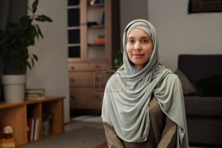 Foto de Retrato mediano de una joven musulmana con hijab sentado en el suelo en la sala de estar en casa mirando a la cámara - Imagen libre de derechos