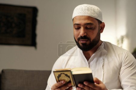 Medium closeup of bearded Muslim man wearing white taqiyah reading holy Quran book, copy space