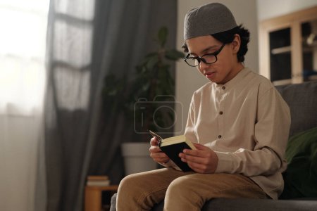 Foto de Mediana toma de niño musulmán con anteojos sentados en el sofá leyendo libro sagrado del Corán, espacio para copiar - Imagen libre de derechos