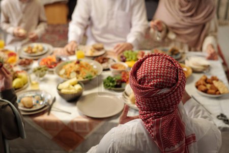 Foto de Vista de ángulo alto enfoque selectivo tiro de hombre musulmán irreconocible usando keffiyeh tener una cena festiva con su familia, espacio de copia - Imagen libre de derechos