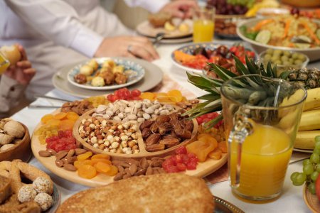 Foto de Captura selectiva de frutas secas dulces y nueces en bandeja de madera en la mesa de comedor festiva, personas musulmanas irreconocibles celebrando Eid Al-Fitr - Imagen libre de derechos