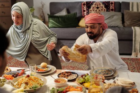 Foto de Hombre musulmán usando keffiyeh arrancando pedazo de pan plano durante la cena festiva con la familia en Eid Al-Fitr - Imagen libre de derechos