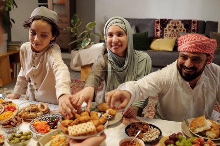 Happy Muslim man, woman and kid taking desserts from plate unrecognizable man holding at family dinner on Uraza Bayram