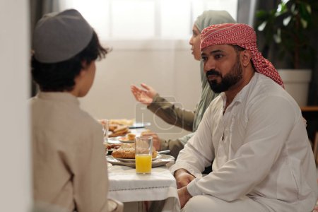 Téléchargez les photos : Mise au point sélective de l'homme musulman portant kandora et keffiyeh assis table à manger festive bavarder avec son neveu pendant le dîner de famille - en image libre de droit