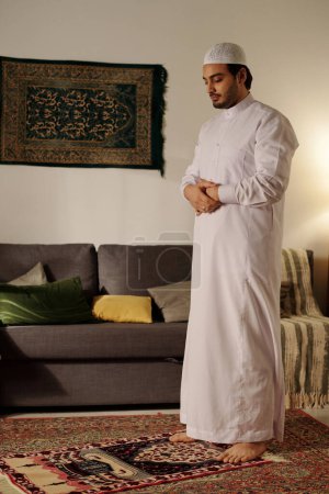 Vertical long shot of young Muslim man in white clothes standing with eyes closed on prayer rug doing namaz in living room