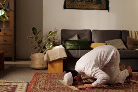 Jeune homme du Moyen-Orient pratiquant le namaz sur un tapis de prière faisant sujud dans le salon à la maison, espace de copie