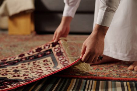 Foto de Medio primer plano de hombre musulmán irreconocible colocando alfombra de oración en el suelo en la sala de estar antes de hacer namaz, espacio de copia - Imagen libre de derechos
