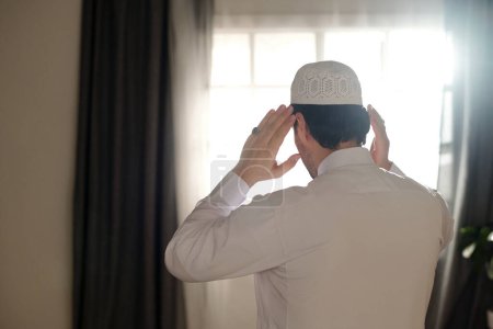 Rear view of unrecognizable young Muslim man wearing white thobe and taqiyah praying salah, copy space