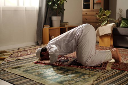 Téléchargez les photos : Jeune homme musulman portant des vêtements blancs faire sajdah tout en pratiquant namaz sur le tapis de prière dans le salon à la maison - en image libre de droit