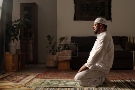 Foto de Vista lateral de hombre musulmán maduro con ropa blanca sentado en la alfombra de oración en la sala de estar haciendo namaz - Imagen libre de derechos