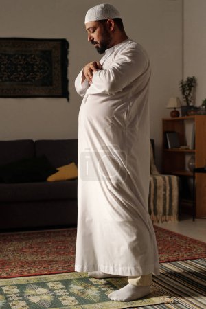 Foto de Tiro completo vertical de hombre musulmán barbudo con toba blanca y taqiyah de pie en la alfombra de oración haciendo namaz en la sala de estar - Imagen libre de derechos
