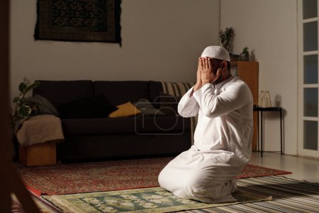 Unrecognizable Muslim man wearing white clothes sitting on rug on floor praying salah in spacious living room, copy space