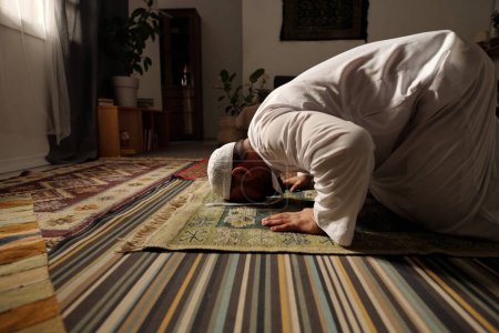 Foto de Vista lateral del hombre musulmán barbudo con ropa blanca rezando salah en la alfombra en la sala de estar haciendo sajdah, espacio de copia - Imagen libre de derechos