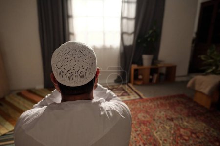 Foto de Vista trasera de un musulmán irreconocible vestido con toba blanca con taqiyah rezando salah en la sala de estar en casa, espacio para copiar - Imagen libre de derechos