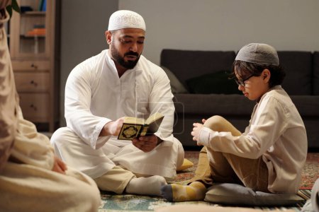 Téléchargez les photos : Homme musulman barbu assis sur le sol dans le salon expliquant la signification de la sourate du Coran à son fils et sa fille lors d'une réunion de famille - en image libre de droit