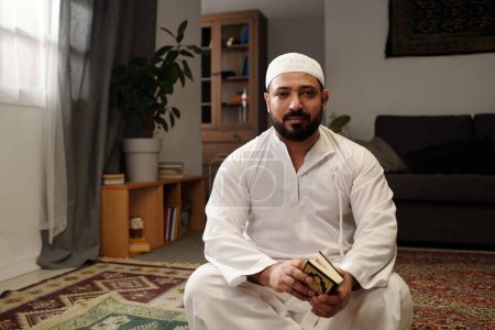 Téléchargez les photos : Portrait de l'homme musulman barbu assis sur le sol dans le salon avec livre du Coran dans les mains regardant la caméra, l'espace de copie - en image libre de droit