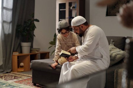 Téléchargez les photos : Homme musulman barbu moderne portant kandora assis sur le canapé dans le salon expliquant la sourate du Coran à son fils, espace de copie - en image libre de droit