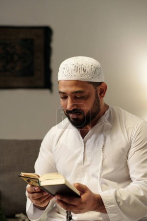 Photo for Vertical medium portrait of bearded Muslim man wearing white clothes holding misbaha reading Quran book - Royalty Free Image