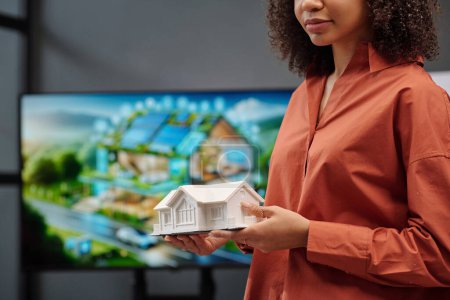 Arquitecta seria en camisa marrón sosteniendo modelo de casa mientras está de pie contra la pantalla con plantilla visual de su proyecto
