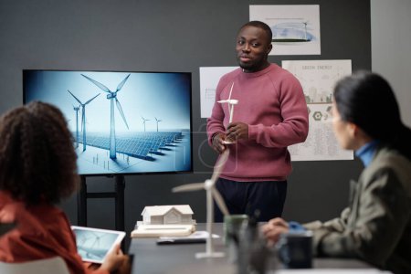 Foto de Joven hombre de negocios con modelo de molino de viento en las manos mirando a una colega mientras está de pie junto al tablero interactivo durante la presentación - Imagen libre de derechos