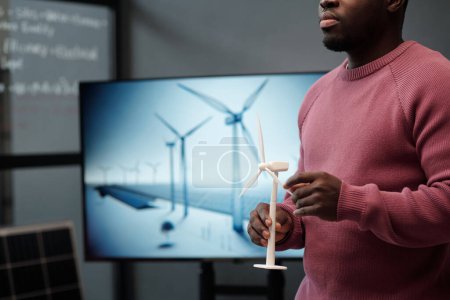 Cropped shot of young male manager with model of windmill in hands standing against interactive board with visual template of his project