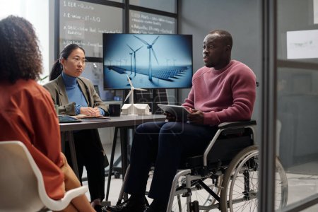 Arquitecto afroamericano masculino con tableta sentado en silla de ruedas delante de colegas femeninas y haciendo presentación de la energía del molino de viento